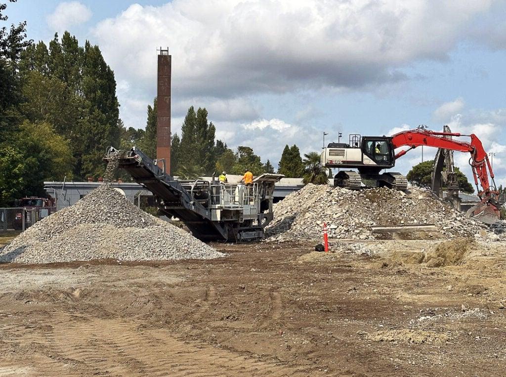 an expanse of concrete leads to two partially demolished buildings - one on each side - each has an excavator doing demolition